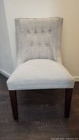Front view of light beige upholstered chair with button-tufted back and dark wooden legs on wood floor.