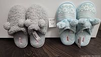 Two pairs of slippers placed side by side against white background and dark floor: grey plush with pom-poms on the left; light blue snowflake knit with fuzzy cuff and pom-poms on the right