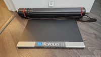 Black extendable plastic drawing tube and closed Itoya Art Profolio portfolio laying flat on floor.
