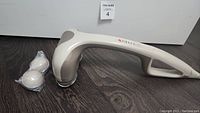 Beige Homedics handheld percussion massager laying flat on dark wood surface with two round additional massage heads wrapped in plastic beside it, showing ergonomic handle and control panel.