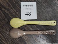 Top view of two stoneware spoons side-by-side with auction card number 48, showing their full length and colors (yellow and light brown).