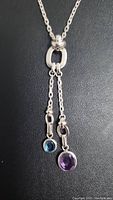 Close-up photo of the necklace's two colored gemstones set in sterling silver, showing blue and purple round gem pendants hanging from a silver loop detail on chain.