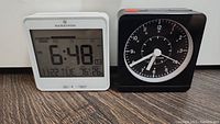 Marathon atomic digital clock in white showing time 6:48 AM, date 11/22 Tuesday, temperature 76°F, and humidity 26% alongside black analog alarm clock with white dial and silver hands.