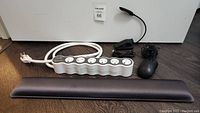 Photo showing all four items laid out: white 360electrical power bar with six outlets, black Targus wired mouse, black rechargeable LED book light, and grey cushioned wrist support pad.