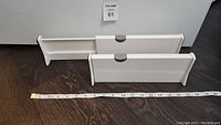 Two white adjustable drawer dividers placed on dark wooden floor, alongside tape measure showing about 17 inches extended length