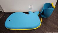 Blue whale-shaped bath kneeler with yellow edges and rinser cup on side