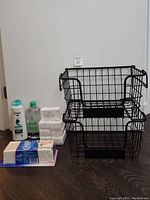 Full view of three stacked black wire baskets with bathroom supplies to the side: shampoo, oil, soap bars, and Q-Tips.