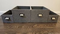 Front view of 4 grey linen look storage boxes, 2 small and 2 medium size, with metal handles and label slots, placed side by side on wood floor.