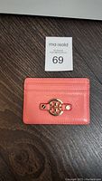 Front view of the coral pink leather card case showing the gold Double T logo hardware on a dark wood surface with a maxsold lot number card.