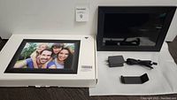 Photo of UCMDA digital photo frame in box along with power adapter and stand parts on white surface.