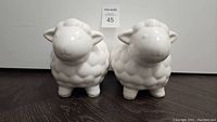 Front view of two white ceramic lamb coin banks side by side on wood surface