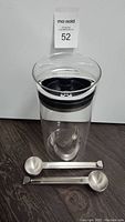 Full front view showing clear glass cylindrical canister with silver metal lid having black seal and two metal scoop spoons placed in front on dark surface.
