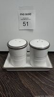 Front view of the white ceramic salt and pepper shakers with holes on top, placed on a rectangular tray with gray rim.