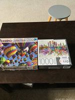 Two puzzle boxes on a dark wooden table, one showing hot air balloons, the other a city skyline.