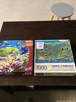 Photo of two 1000-piece puzzles on a dark wood table with a small round stool in the background. Puzzles include an underwater coral reef scene and a coastal town with hot air balloons and whales.