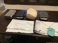 Three dress up pocketbooks displayed on dark table with dust bags underneath each one including Jimmy Choo and Maxima Marina branded dust bags plus Tiffany & Co. pouch