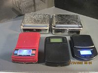 Five items arranged in two rows. Two metal mini scales with engraved lids in back row. Three items in front row: red mini scale with illuminated LCD, black mini scale with display, and a black manual tobacco roller with chrome roller mechanism.