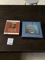 Two audio CD sets on a dark wooden table; Brian Tracy's 'The Psychology of Selling' in dark blue, Earl Nightingale's 'Lead the Field' in red and white case.