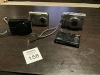 Four digital devices on table: Fujifilm FinePix cameras, HP Photosmart M437 camera, Max V16 MP6 media player, some with wrist straps.