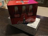 Red Coca Cola collector tin on dark table, showing front with three vintage glass bottles and Coca Cola logo.