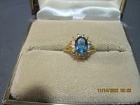 Front view of the gold ring with oval blue stone and halo of small clear stones in a beige jewelry box.