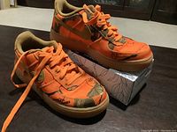 Pair of Nike Air Force 1 sneakers resting on a box showing the top and side with bright orange and Real Tree camo fabric, tan swoosh and gum sole.