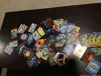 Overview of assorted Pokémon and Digimon trading cards spread out on a dark table, showing various designs including colorful card backs and fronts with character images.