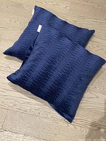Two square blue decorative cushions with textured horizontal stripe pattern, arranged overlapping on a light wood floor.