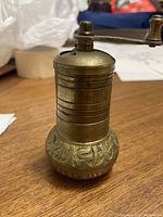 Brass pepper grinder standing upright on wooden surface showing engraved lower body and smooth top with crank handle.