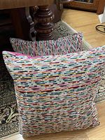 Two square pillows with multi-colour linear patterns on the front, resting on a patterned rug with visible pink velveteen on one pillow's back.