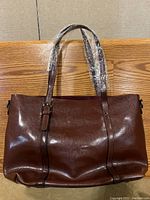 Front view of brown leather handbag showing two handles wrapped in plastic and structured body.