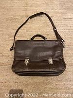 Front view of the brown leather briefcase with shoulder strap and dual buckle closures.