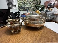 Photo showing two silver plated serving vessels: a larger covered serving dish with a ring handle on the lid and three feet, and a smaller covered sugar or jam dish with a knob handle and side handle.