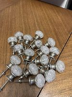 Seventeen clear glass decorative knobs with floral texture on metal bases arranged on wooden surface.