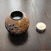Round wood candle holder with metal leaf accents and tea light candle beside it on a dark wood surface.