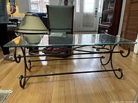 Side angle of glass and wrought iron coffee table on wooden floor with a lamp and furniture in background
