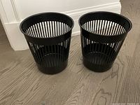 Two black Ikea plastic trash bins with vertical slats, positioned side by side on wooden floor.