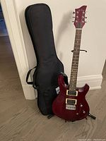 Photo shows red Jay Turner electric guitar leaning on a black metal stand, next to a black padded soft carrying bag upright against the wall.