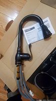 Moen brand kitchen faucet with high-arc curved spout and single lever control, showing attached supply lines and instruction manual on cardboard base.