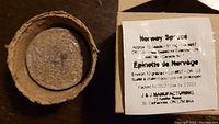 Close-up image of a biodegradable peat pot with soil and a sealed white packet labeled "Norway Spruce" containing about 10 seeds with lot number and seed weight info.