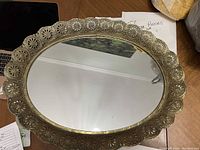 Top view of the oval mirrored tray showing the brass-colored metal frame with open circular pattern around the edges and the reflective mirror surface in the center.