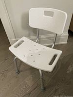 Photo of white plastic shower chair with backrest and adjustable aluminum legs with rubber non-skid tips on hardwood floor.