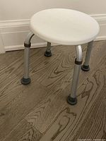 Photo of a round white shower stool with four adjustable aluminum legs ending in black rubber non-slip tips on wood floor.