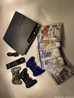 Sony PS3 Slim console with 3 wired controllers in black, blue, and camouflage, 5 sports video games including NHL and MLB titles, and power cable.