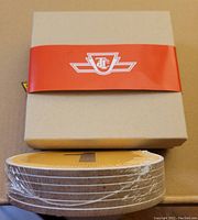 Stack of six round yellow coasters with white edges sealed in shrink wrap in a beige box with a red TTC band.