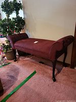 Front and side view of the upholstered wooden bench showing burgundy fabric and curved wooden legs with rolled arms at each end.