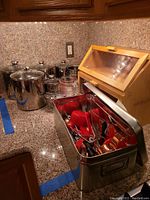 Kitchen countertop with canisters and bread box at back, open metal box with red and metal kitchen utensils and measuring tools in front.