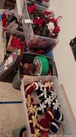 5 plastic tubs on floor filled with assorted Christmas floral and decorative items including floral picks, trays, garlands, and ornaments.