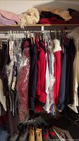 Rack of hanging coats, jackets, and other outerwear with some items in plastic covers, folded textiles on upper shelf.