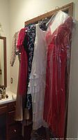 Photo showing five dresses hanging on a wooden hanger rack, featuring a long red gown, a white ruffle dress, a blue floral print dress, a leopard print dress, and a beige patterned dress.